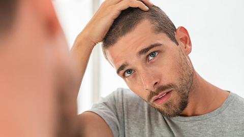 Ein junger Mann sucht im Spiegel nach grauen Haaren an seinem Kopf - Foto: iStock/Ridofranz