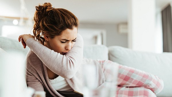 Eine erkältete Frau liegt auf dem Sofa und hustet in ihre Armbeuge - Foto: iStock_Drazen Zigic