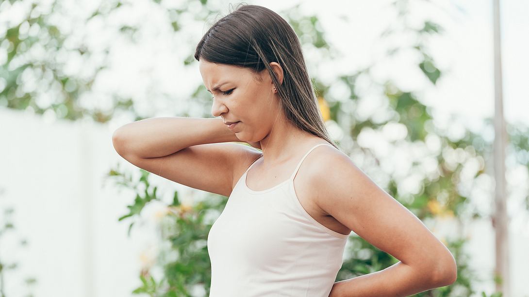 Junge Frau im Top fasst sich mit schmerzverzerrtem Gesicht an Rücken und Nacken - Foto: iStock/klebercordeiro