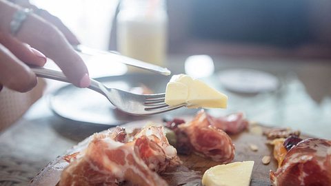 Eine Person nimmt mit einer Gabel ein Stück Käse von einem Brett, auf dem Salami- und Käsestücke liegen - Foto: iStock/webphotographeer