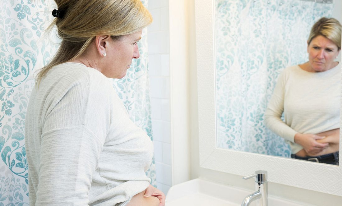 Eine Frau im mittleren Alter steht vor einem Spiegel und betrachtet ihren Bauch