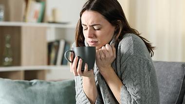 Eine Frau sitzt krank auf dem Sofa und hält eine Tasse in der Hand - Foto:  iStock/AntonioGuillem