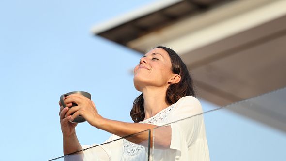 Frau steht auf dem Balkon und genießt die Sonne. - Foto: iStock/Pheelings Media