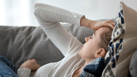Eine Frau liegt krank auf der Couch und hält sich den Kopf - Foto: iStock/fizkes