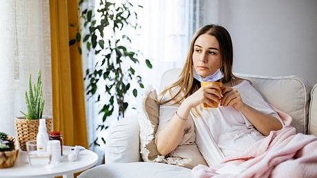 Eine Frau sitzt mit Maske und Tee auf dem Sofa - Foto: iStock_ozgurcankaya