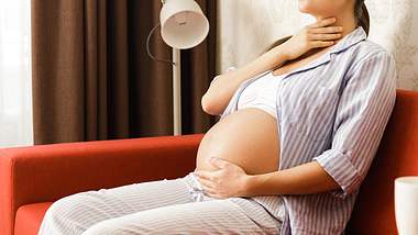 Schwangere Frau sitzt auf einem roten Sofa und fasst sich an den Hals - Foto: iStock/Jun