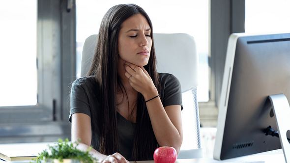 Junge Frau sitzt am Schreibtisch im Büro und fasst an den Hals, das Gesicht schmerzverzerrt - Foto: istock/﻿nensuria