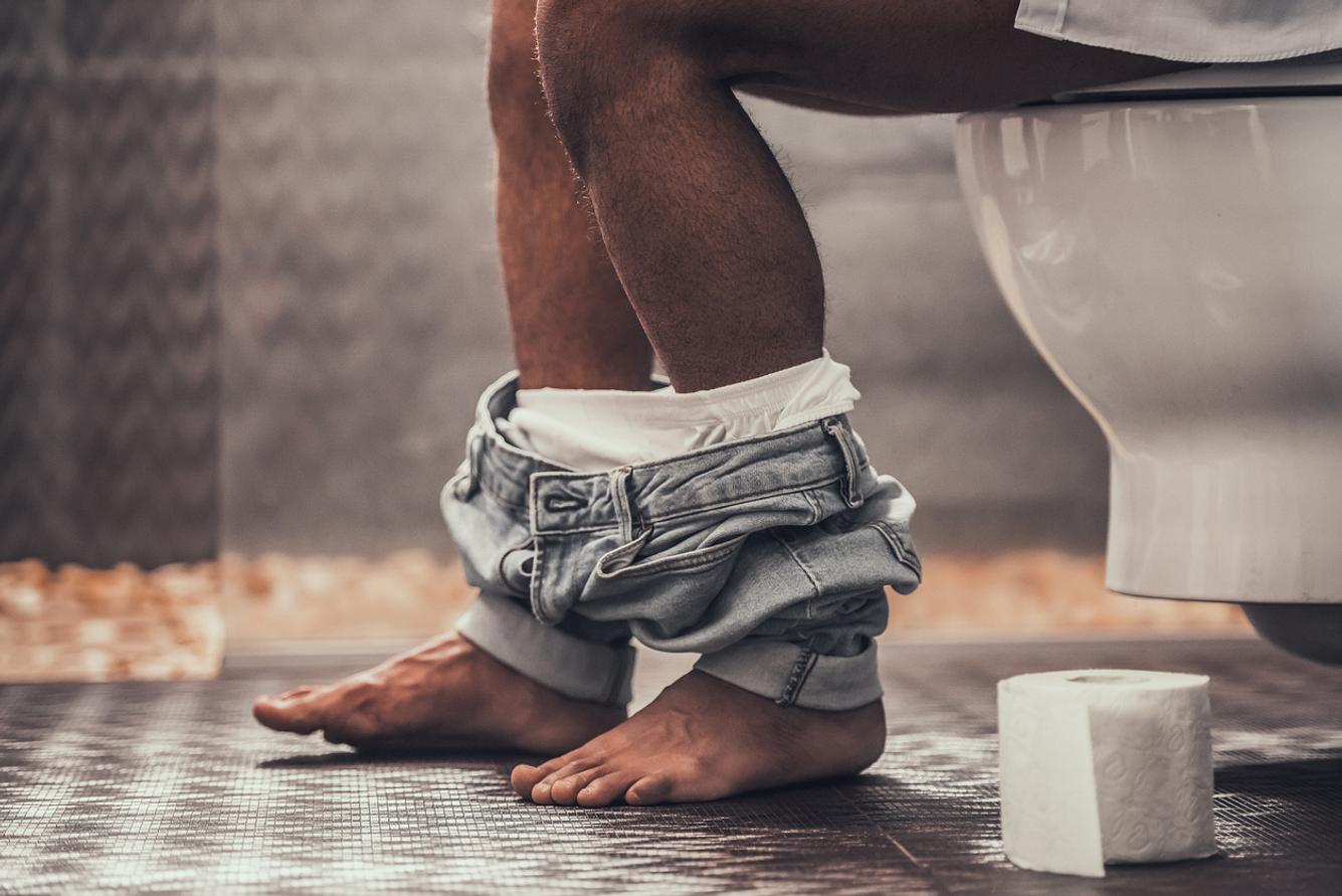 Beine eines Mannes auf der Toilette mit Hose um die Knöchel