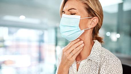 Frau mit Mund-Nasen-Schutz hält sich den Hals - Foto: iStock/PeopleImages