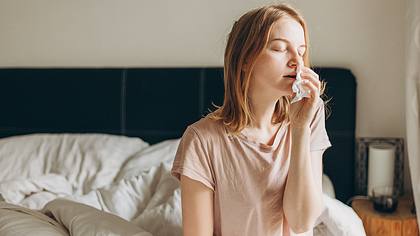 Eine Frau sitzt im Bett und putzt sich die Nase - Foto: iStock/Mariia Skovpen