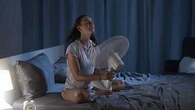 Frau sitzt nachts mit Ventilator im Bett wegen Hitzewallungen - Foto: iStock/cyano66