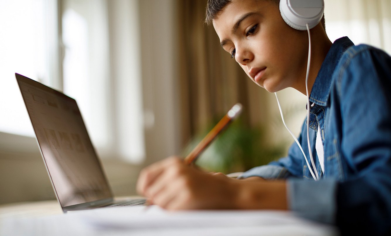 Junge arbeitet am Laptop, hört nebenbei Musik und schreibt etwas mit einem Stift