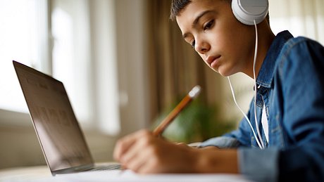 Junge arbeitet am Laptop, hört nebenbei Musik und schreibt etwas mit einem Stift - Foto: istock_damircudic