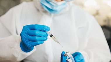 Arzt in Schutzkleidung zieht Impfserum in eine Spritze - Foto: iStock-1223925413 bojanstory