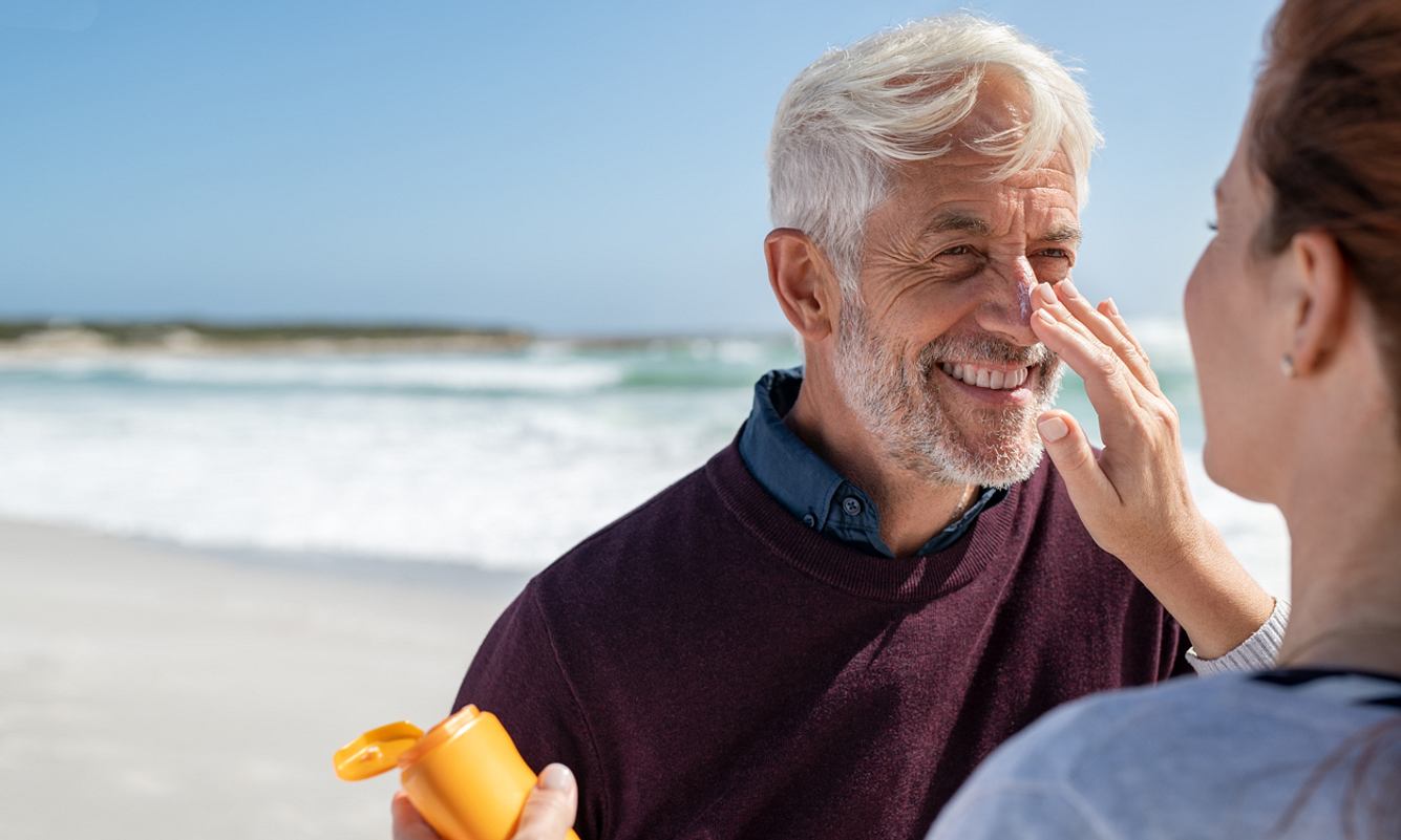 Warum altert Haut schneller Älterer Mann cremt sich am Strand die Nase mit Sonnenmilch ein