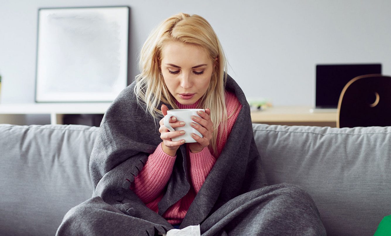 Junge Frau sitzt mit Grippe sitzt mit einer Tasse Tee auf dem Sofa und leidet unter Reizhusten Symptomen