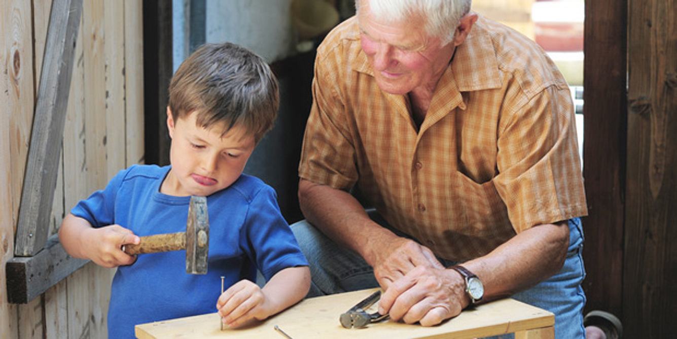 Ein Junge schlägt einen Nagel ins Holz und sein Großvater passt auf