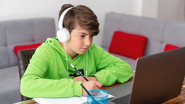 Junge in grünem Hoodie und mit Kopfhörern sitzt zu Hause am Holztisch vor einem Laptop - Foto: iStock-1216236928 SonerCdem