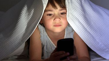 Ein Kind schaut unter der Decke auf sein Smartphone - Foto: iStock