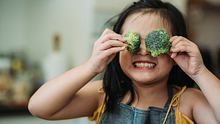 Asiatische chinesische weibliche Kind mit Brokkoliröschen in der Hand, die sie sich mit lächelndem Gesicht vor die Augen hält - Foto: iStock/Marcus Chung
