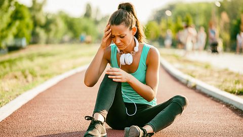 Eine Frau sitzt in Sportkleidung auf einer Laufbahn und hält sich den Kopf - Foto: iStock/Photodjo