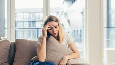 Eine Frau sitzt mit Kopfschmerzen auf einem Sofa - Foto: iStock/Liubomyr Vorona