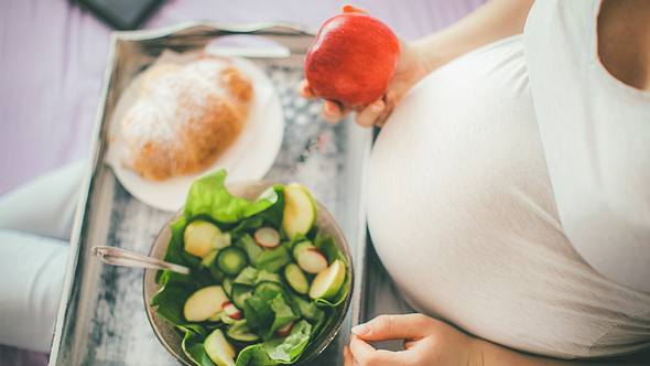 Bauch einer Schwangeren von oben, vor ihr ein Tablett mit Salat und Croissant; in der Hand hält sie einen Apfel - Foto: iStock-636752590 Pekic