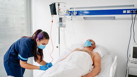 Patient mit Schutzmaske im Krankenbett; Schwester verabreicht Infusion - Foto: istock/Phynart Studio