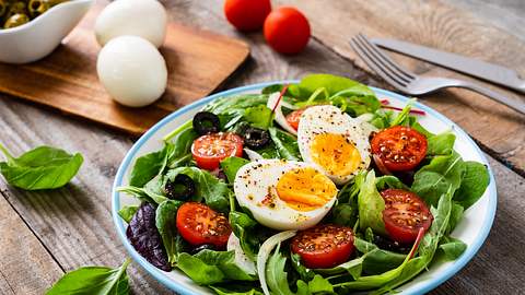 Teller mit Salat, Tomaten und zwei halben Eiern auf einem Holztisch; im Hintergrund Besteck und ein Brettchen mit Eiern - Foto: iStock-1194610986 gbh007