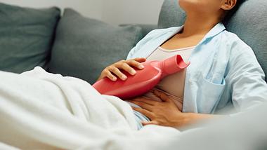 Frau mit Wärmflasche auf dem Sofa - Foto: iStock/Drazen Zigic