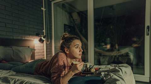 Eine Frau liegt mit Essen im Bett und schaut Fernsehen - Foto: iStock/AleksandarNakic