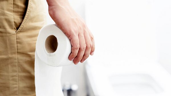 Hosenbein eines Mannes, Hand hält Toilettenpapierrolle, im Hintergrund eine Toilette - Foto:  iStock/Rattankun Thongbun