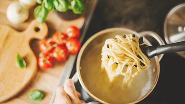 Löffel mit Pasta wird aus einem Topf gehoben, daneben  ein Holzbrett, Tomaten, Basilikum und Knoblauch - Foto: iStock/nerudol