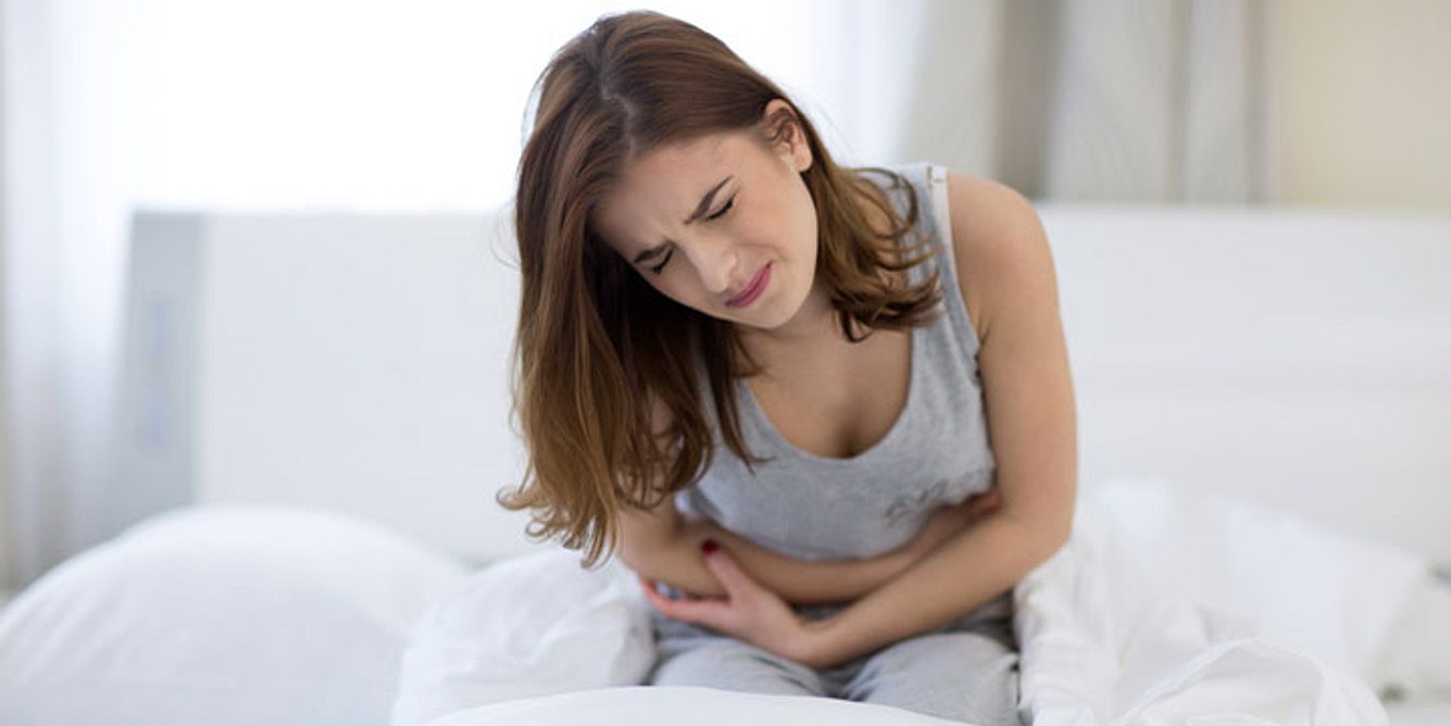 Eine Frau sitzt mit Oberbauchschmerzen auf einem Bett