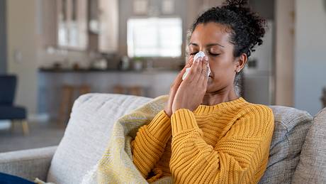 Frau in gelbem Pulli sitzt mit einer Decke auf dem Sofa und schnäuzt sich die Nase - Foto: istock / Ridofranz