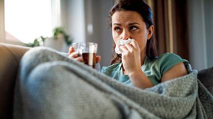 Eine Frau sitzt auf der Couch mit einer Teetasse und einer Decke und putzt sich die Nase - Foto: iStock/damircudic