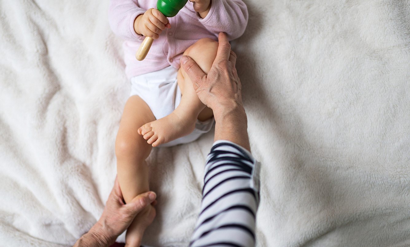 Baby liegt auf einer Decke mit Rassel in der Hand und wird gerade an den Beinen osteopathisch behandelt