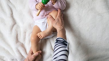 Baby liegt auf einer Decke mit Rassel in der Hand und wird gerade an den Beinen osteopathisch behandelt - Foto: iStock/sanyasm