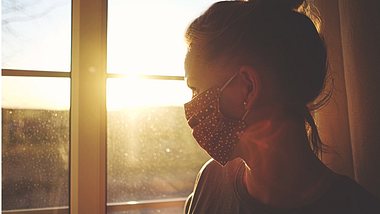 Eine Frau mit Maske schaut aus dem Fenster - Foto: iStock/Rike_