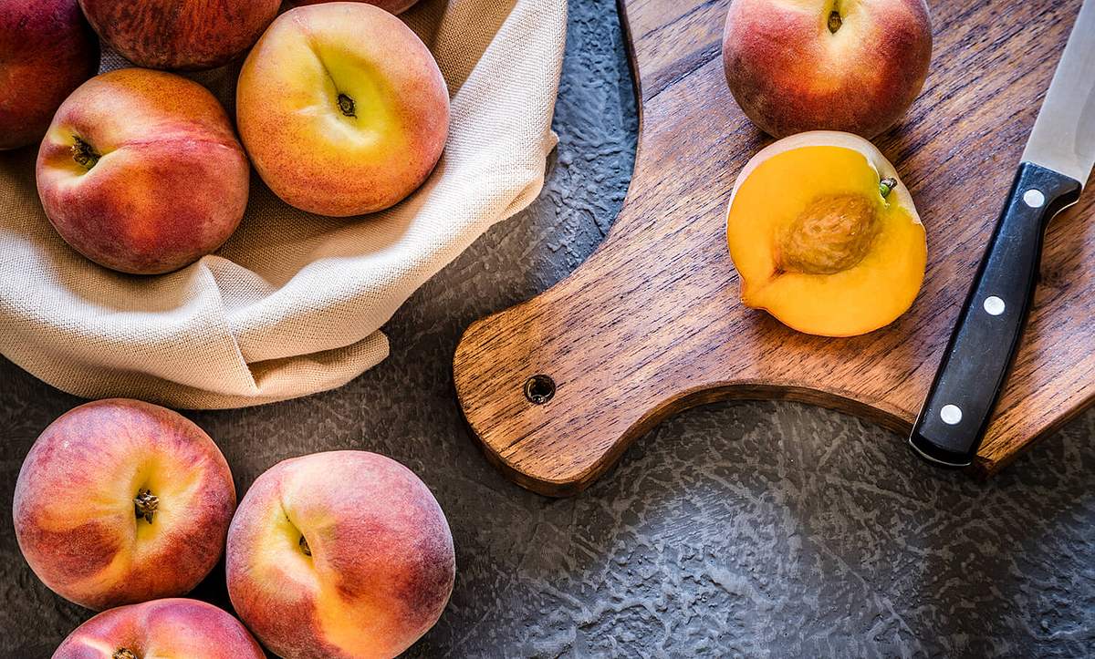 Pfirsich liegt aufgeschnitten mit einem Messer auf einem Brett, daneben liegen weitere Pfirsiche auf dem Tisch oder in einem Beutel