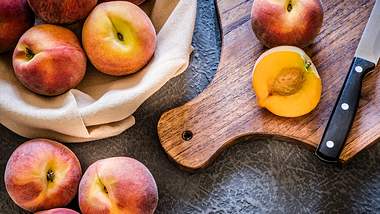 Pfirsich liegt aufgeschnitten mit einem Messer auf einem Brett, daneben liegen weitere Pfirsiche auf dem Tisch oder in einem Beutel - Foto: iStock/carlosgaw