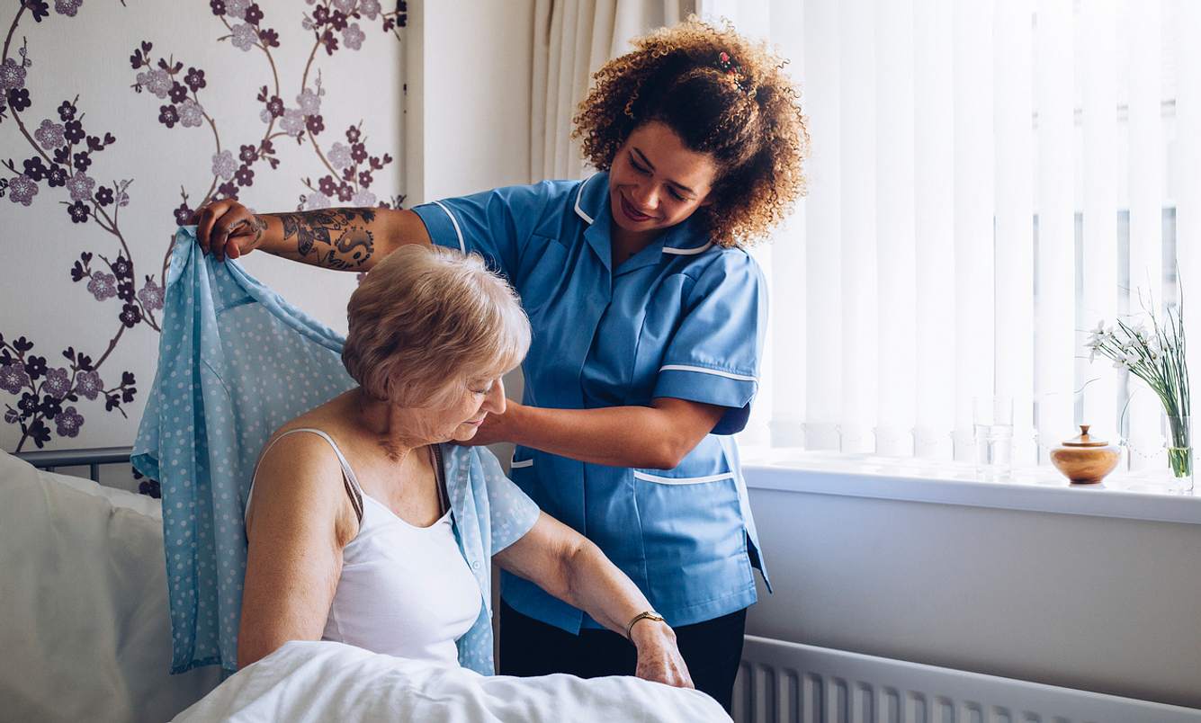 Pflegekraft hilft Patientin auf dem Bett aufzustehen und die Jacke anzuziehen