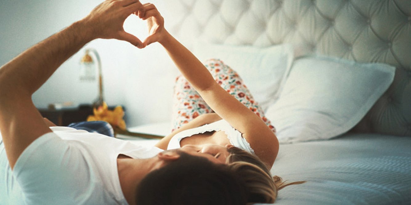 Ein Pärchen liegt im Bett und formt mit den Fingern ein Herz