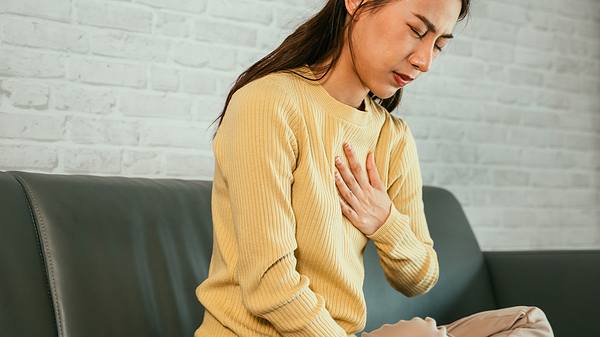 Eine Frau sitzt auf dem Sofa und fasst sich ans Brustbein - Foto: iStock/AsiaVision