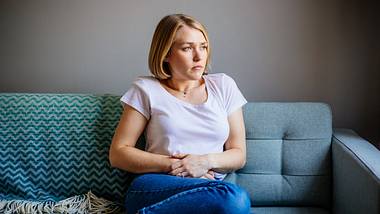 Frau mit Bauchschmerzen sitzt auf dem Sofa und hält sich den Bauch - Foto: iStock/agrobacter