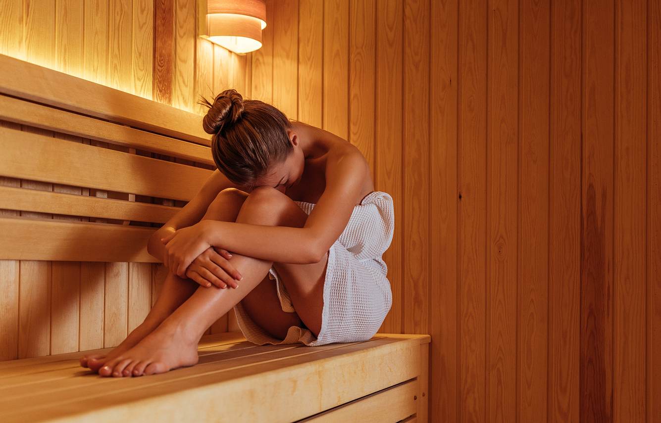 Eine Frau sitzt, mit Handtuch bekleidet, in der Sauna und blickt nach unten