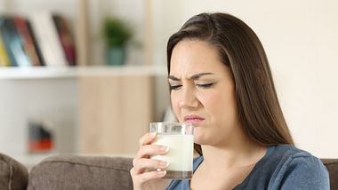 Junge Frau sitzt auf dem Sofa und riecht mit angeekeltem Gesicht an einem Glas Milch - Foto: iStock/AntonioGuillem