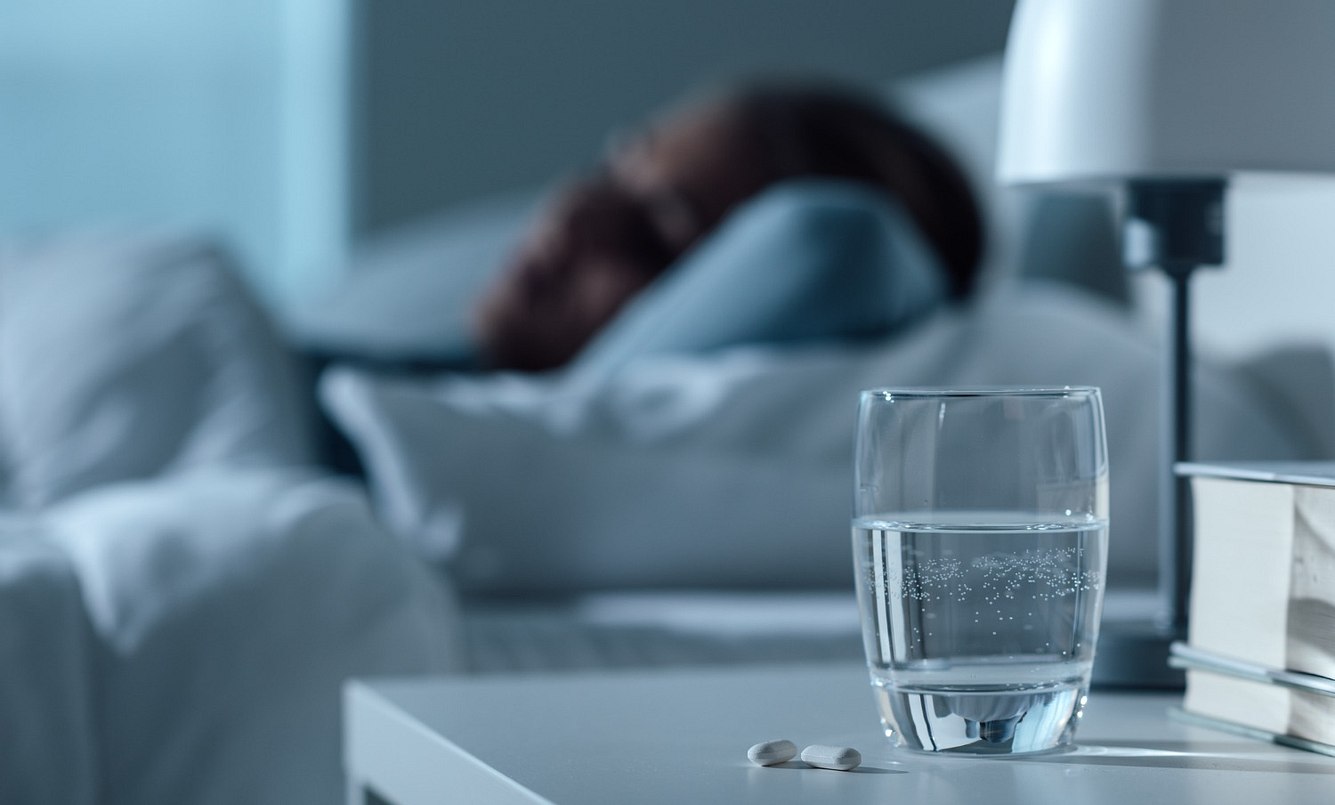 Ein Glas Wasser, daneben eine Schlaftablette auf einem Nachtisch, im Hintergrund eine schlafende Frau