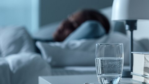 Ein Glas Wasser, daneben eine Schlaftablette auf einem Nachtisch, im Hintergrund eine schlafende Frau - Foto: iStock_demaerre