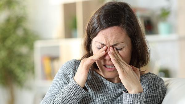 Frau im grauen Pulli sitzt auf dem Sofa und reibt sich die Augen, das Gesicht schmerzverzerrt - Foto: istock / Pheelings Media